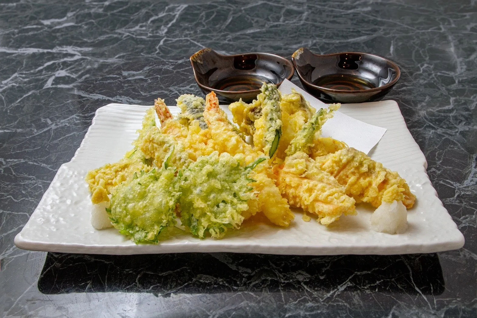 天ぷらの盛り合わせ。エビ、野菜など様々な具材が揚げられ、白い皿に盛り付けられている。テーブルには、天つゆ用の器が置かれ、大根おろしも添えられている。