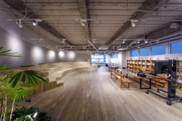 モダンなデザインのオフィスラウンジ。木製の床、階段状の座席、植物、本棚、窓が特徴です。