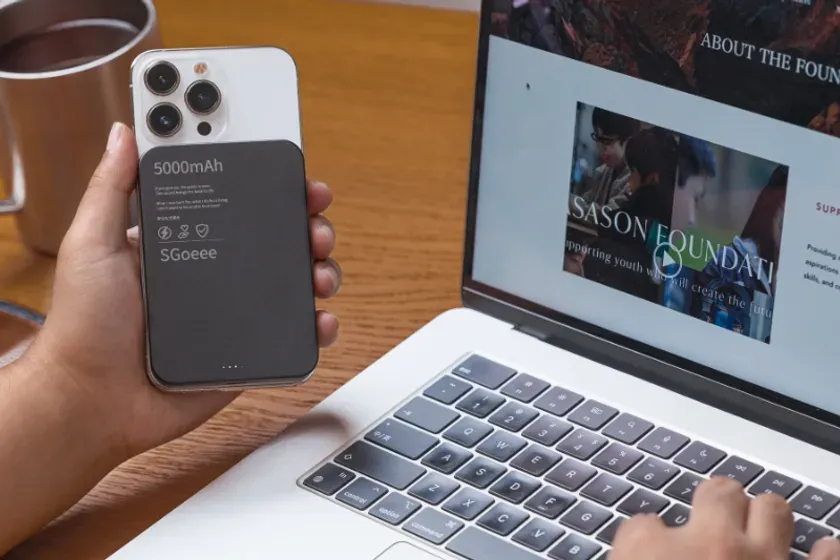 デスクでノートパソコンを使用しながら、iPhoneに5000mAhのSGOeee製モバイルバッテリーを装着している様子です。ノートパソコンの画面には「SASAN FOUNDATION」のウェブサイトが表示されており、現代の作業環境を示しています。