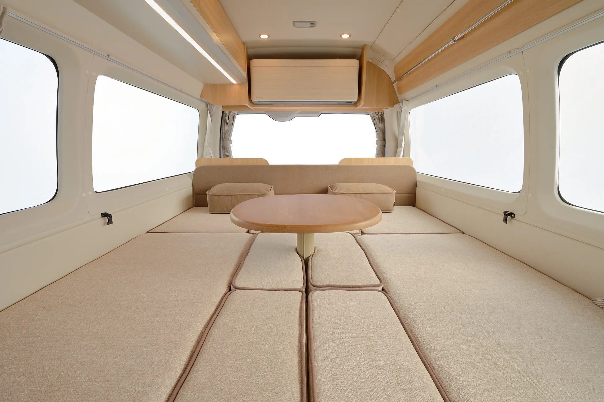 明るいベージュと木目を基調としたキャンピングカーの車内です。中央には円形テーブルがあり、クッションは座席としてもベッドとしても利用できる多機能な空間を演出しています。頭上にはエアコンと照明が備わり、快適な旅を予感させます。