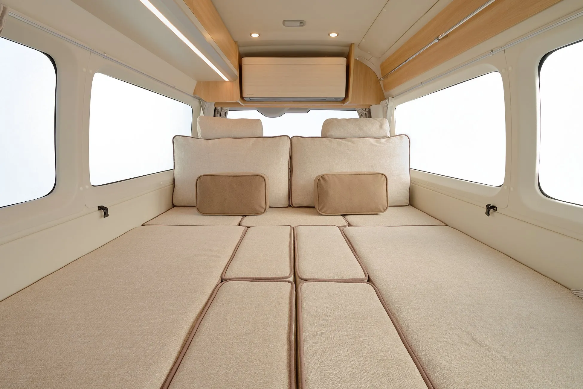 キャンピングカーの車内を写した画像で、広々としたベッドスペースが特徴です。複数のクッションが並べられ、上部にはエアコンも完備されており、快適な車中泊が楽しめそうな内装です。