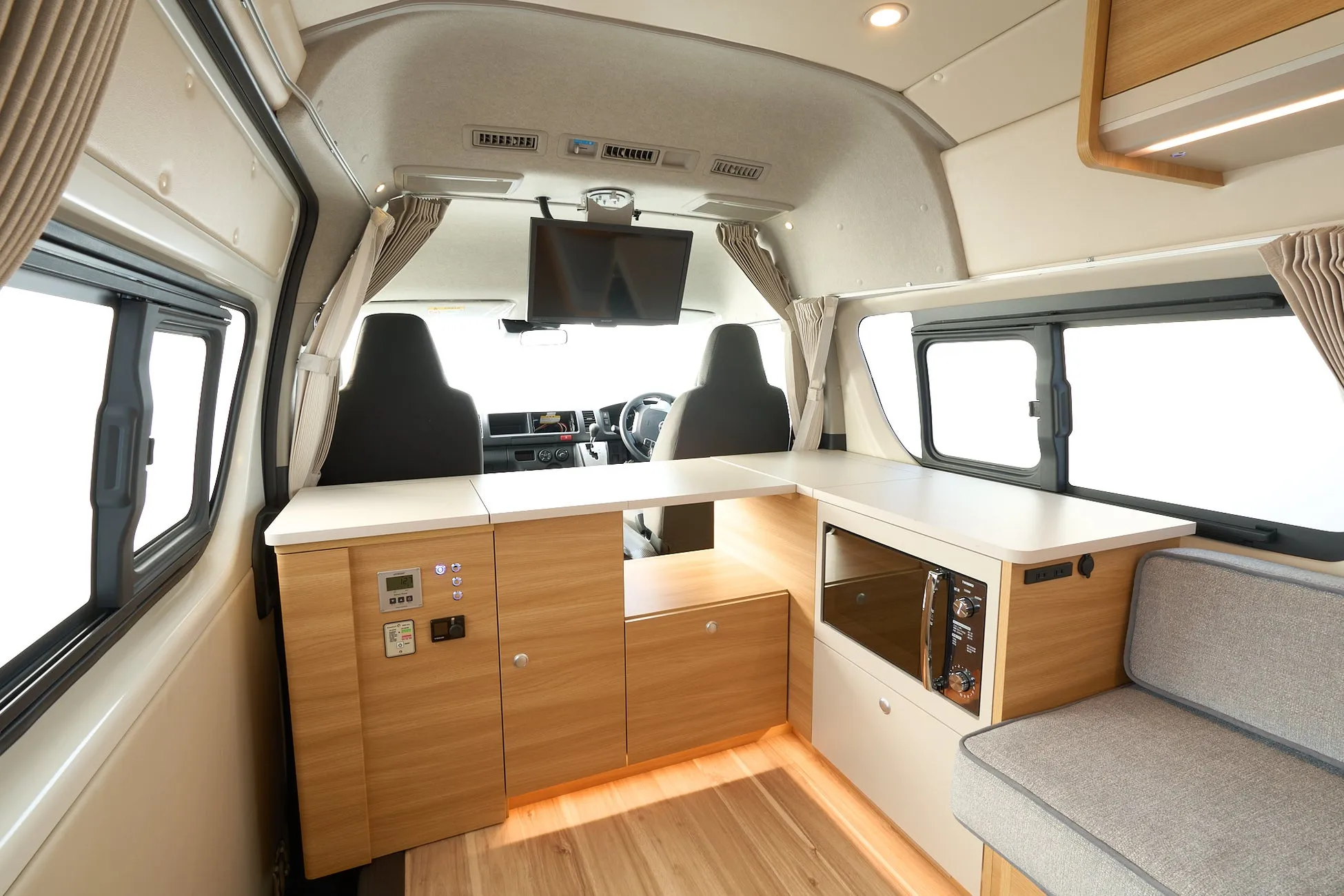 木目調と白を基調としたモダンなキャンピングカーの車内です。カウンター、収納、電子レンジ、ソファ、テレビが完備され、快適な車中泊やバンライフに適した機能的な空間が広がっています。