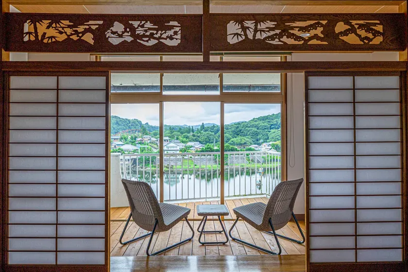和風の部屋から見える風景。窓の外には水辺と緑豊かな景色が広がり、2つの椅子とテーブルが置かれている。
