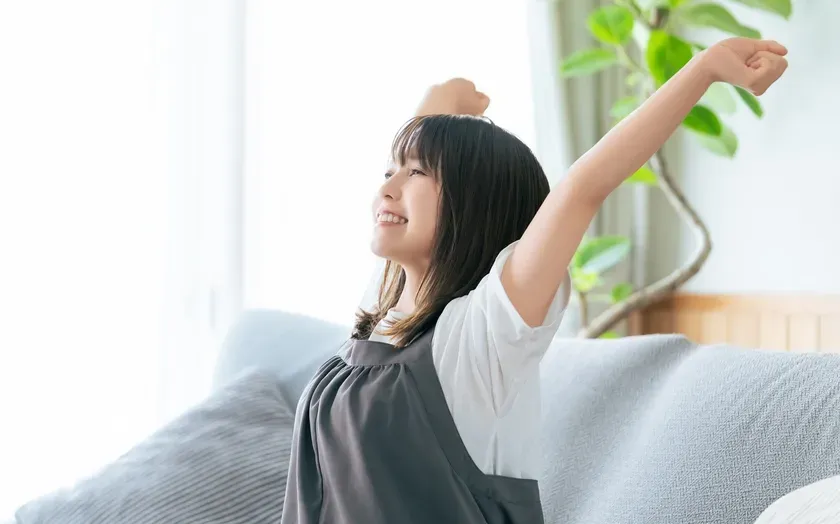 女性がソファに座って伸びをしている様子を写した写真です。彼女は笑顔で、リラックスした様子です。背景には窓と植物があり、穏やかな雰囲気です。
