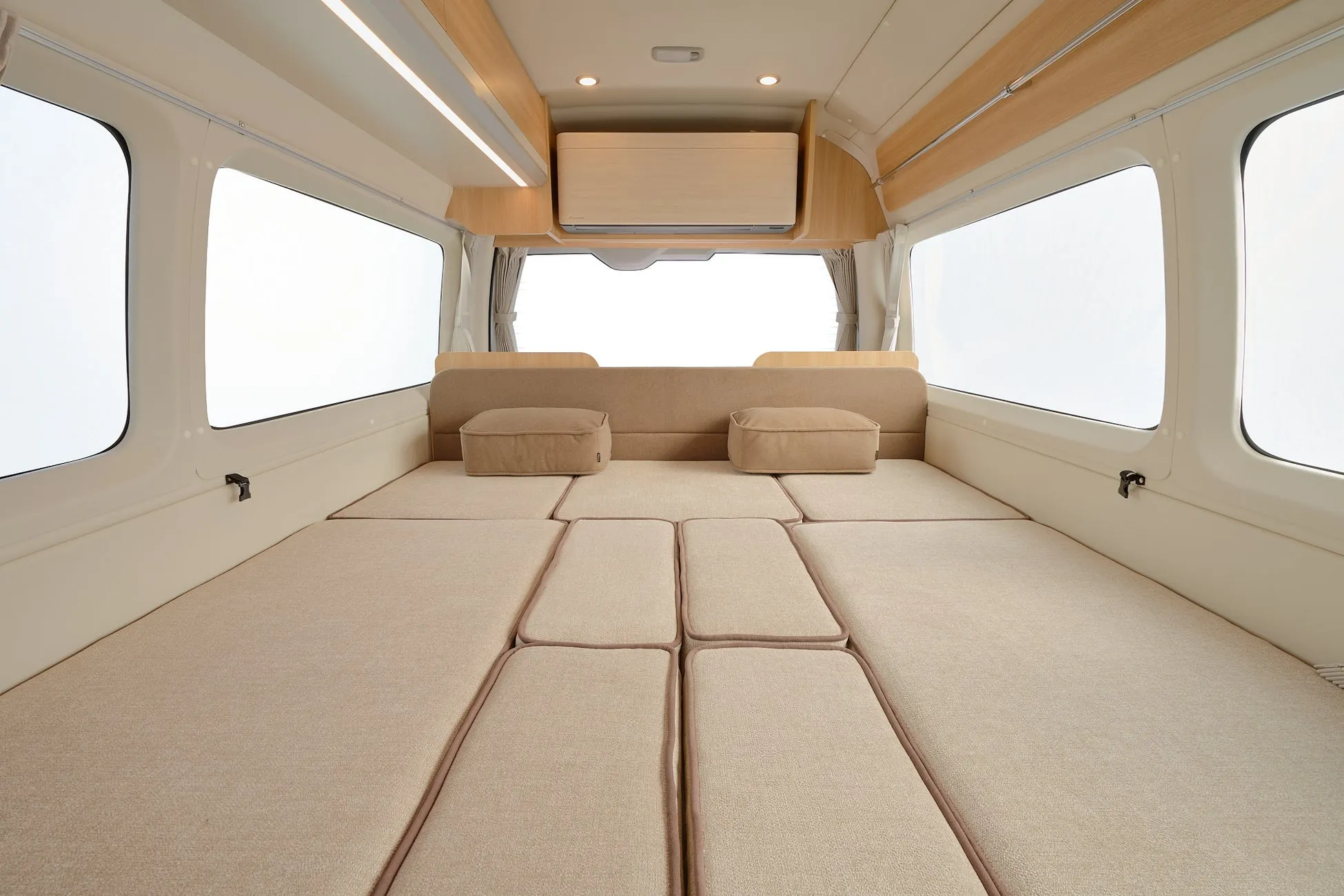 広々としたキャンピングカーの車内を捉えた画像。ベージュのクッションが敷き詰められ、快適なベッドスペースが作られている。両サイドには大きな窓があり、エアコンも完備されたモダンで機能的な空間が広がっている。