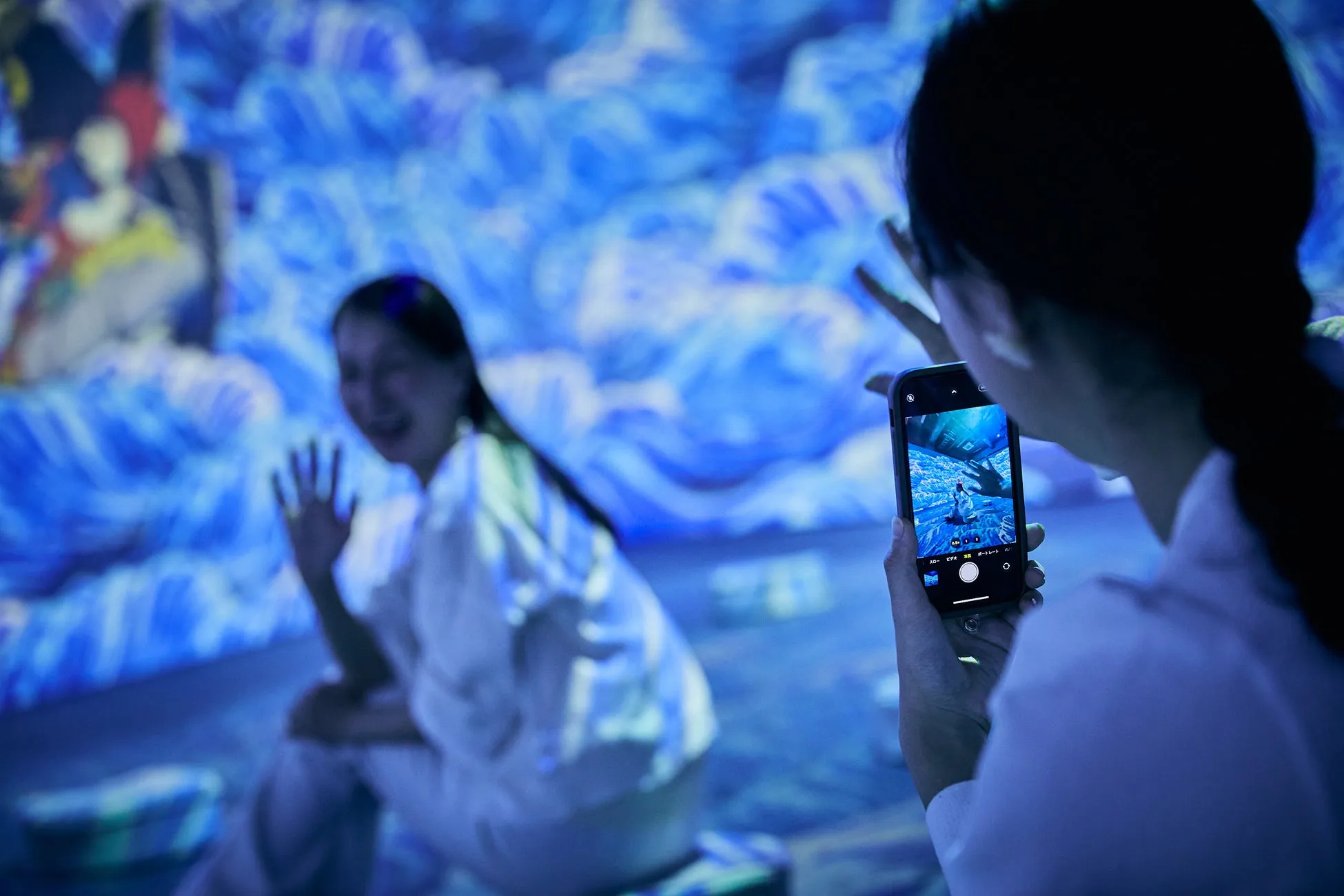 青いデジタルアート空間で、スマートフォンを構えた女性が、波のような映像を背景に笑顔で手を振る別の女性を撮影している。没入型アート体験の様子を捉えた一枚。