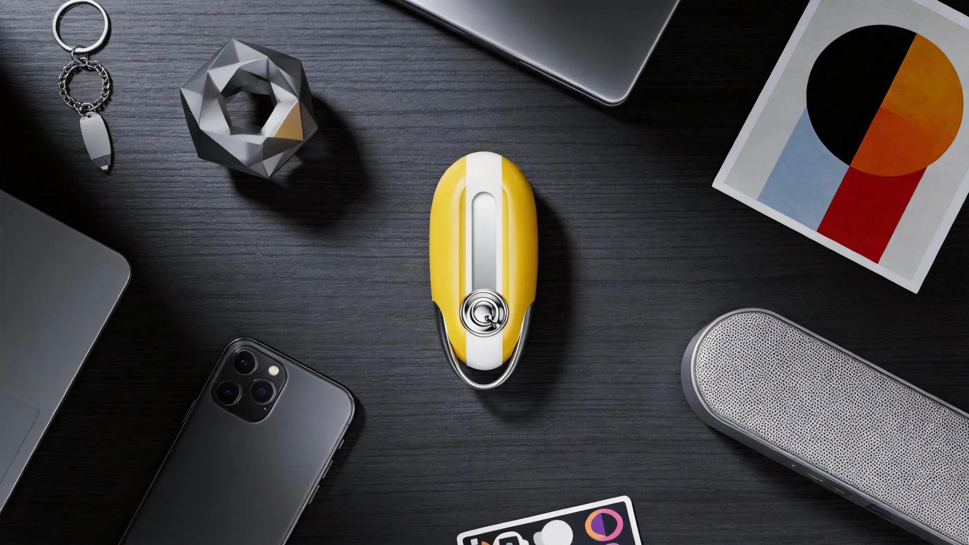 黒い木製のテーブルの上に、キーホルダー、幾何学的なオブジェクト、ノートパソコン、携帯電話、スピーカー、アートワーク、車のキーが配置されています。