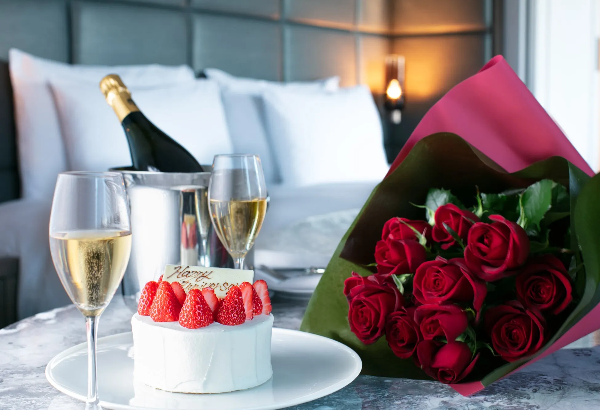ベッドルームのテーブルに、シャンパン、グラス、イチゴのケーキ、バラの花束が置かれたロマンチックなシーン。