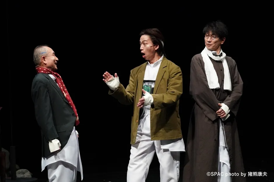 舞台に立つ3人の俳優の画像。真ん中の俳優は口を開け、両手を広げ、右側の俳優は眼鏡をかけ、白いスカーフを巻いている。左側の俳優は笑顔で赤いスカーフを巻いている。