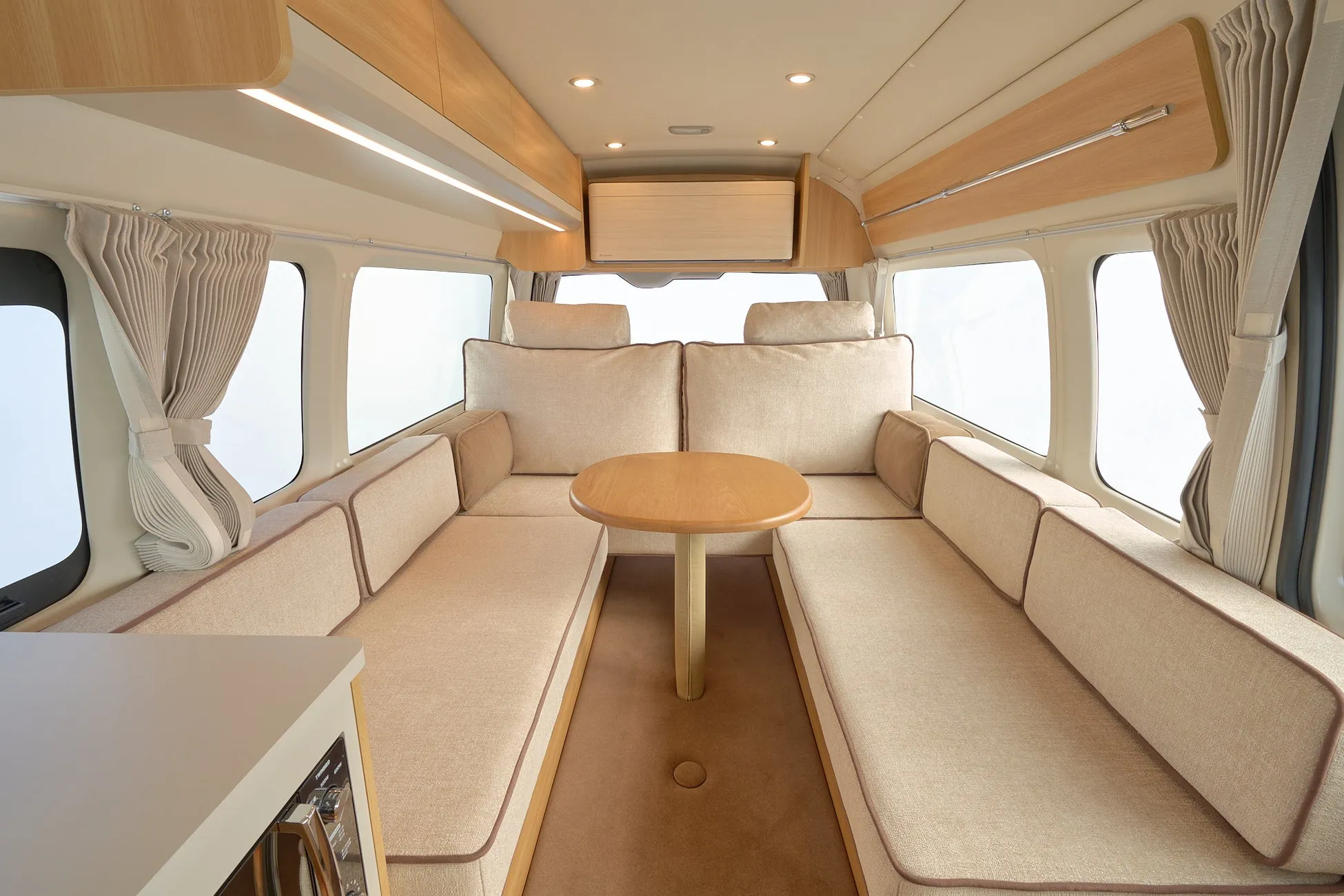 ベージュと木目調で統一された、広々としたキャンピングカーの室内です。中央には丸いテーブルがあり、両サイドにはゆったりとしたソファ席が配置されており、快適な空間が広がっています。窓にはカーテンも備え付けられています。