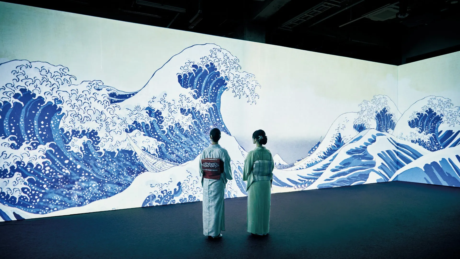 葛飾北斎の『神奈川沖浪裏』の展示を鑑賞する2人の女性の姿を捉えた写真。女性たちは着物を着ており、壁に大きく投影された波の絵を眺めている。