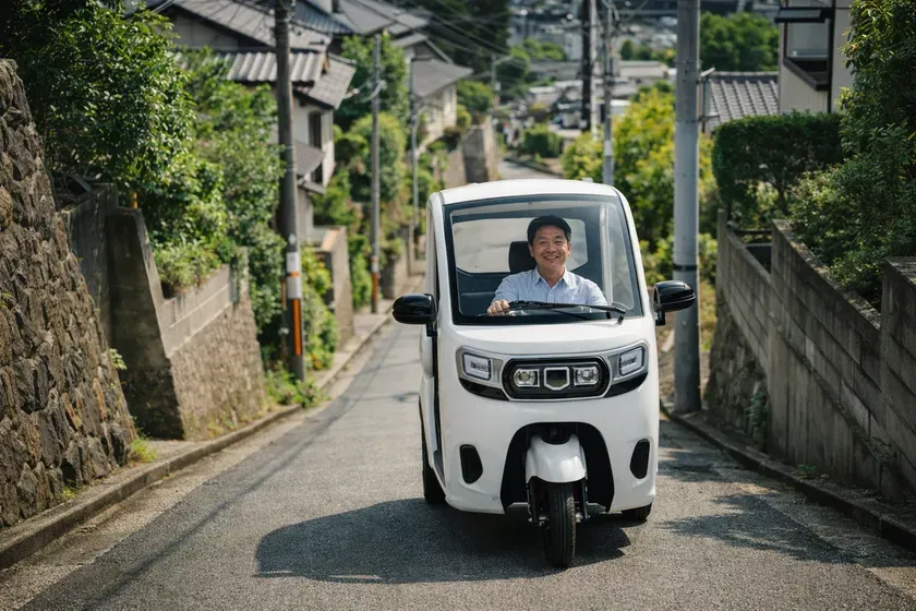 白い三輪の電気自動車に乗って、笑顔で運転する男性の写真。背景には住宅街の風景が広がっています。