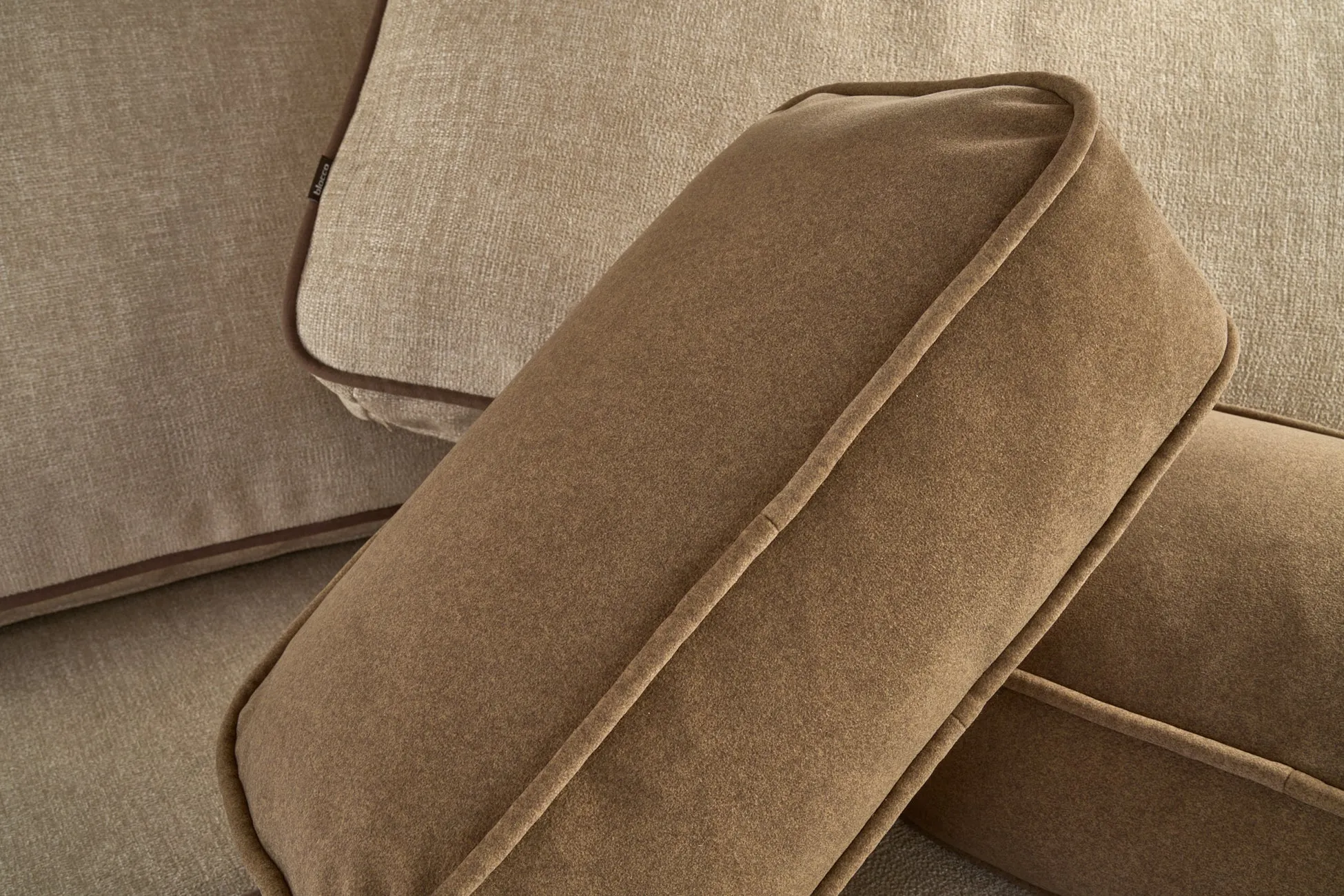 異なる色合いと質感を持つクッションが複数写っています。手前のクッションは滑らかな茶色でパイピングが施されており、奥のクッションはベージュの布地で縁取りがあります。