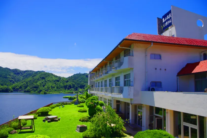 青空の下、湖畔に建つホテルの風景です。ホテルは白壁で、赤い屋根が特徴的です。ホテルの前には緑の芝生が広がり、湖と山々が見えます。