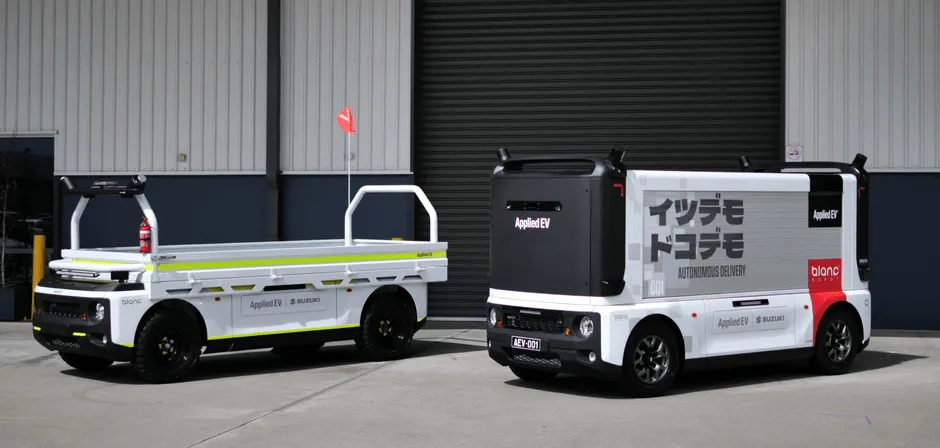 2台の白い電気自動車が並んで駐車されている写真。左側の車両は荷台付きで、右側の車両は箱型の構造をしている。どちらの車両にも「Applied EV」のロゴと「SUZUKI」の文字が見える。