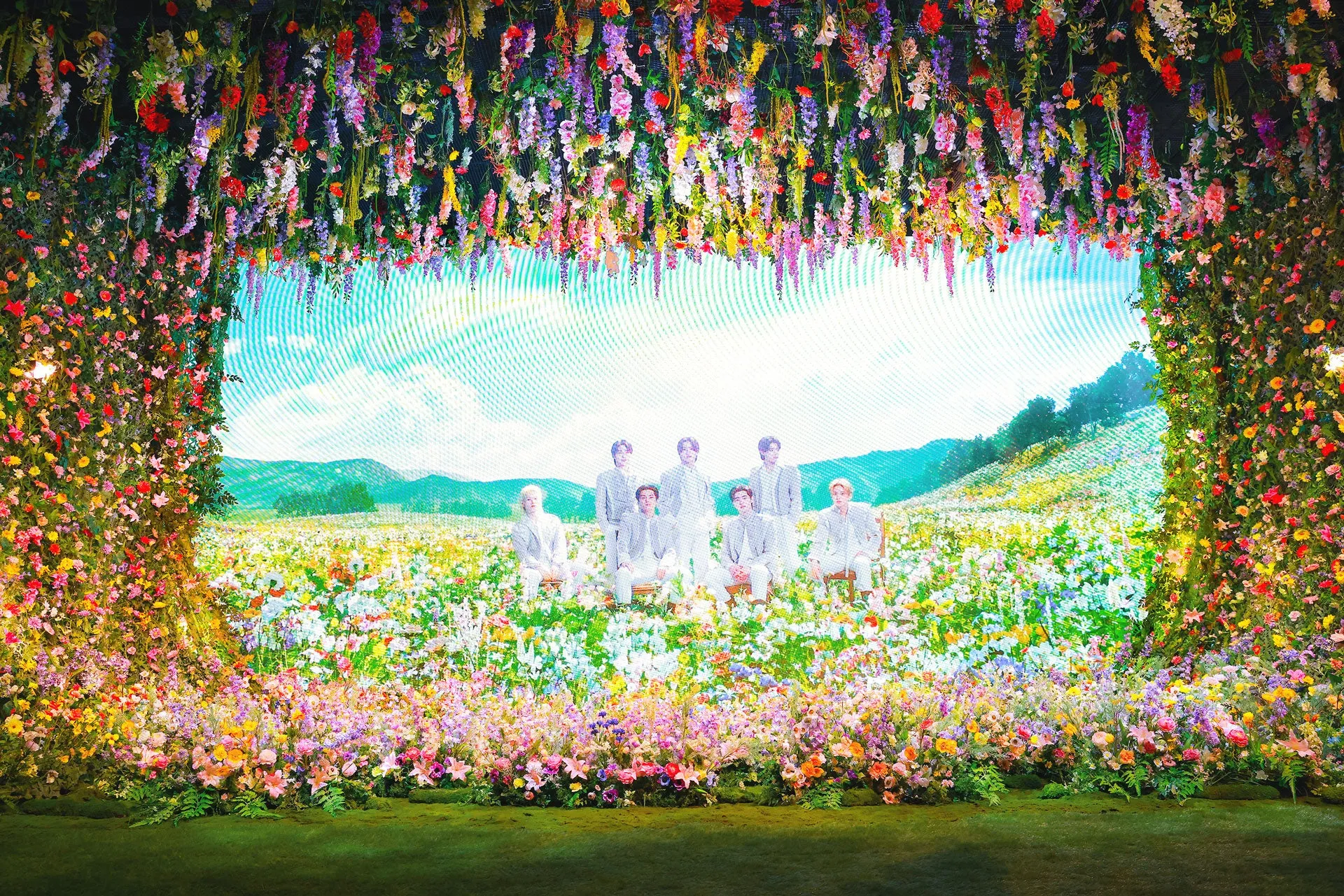 花で飾られたステージでポーズをとる7人の男性のグループの写真。彼らは白いスーツを着ており、花で飾られた環境の中にいます。