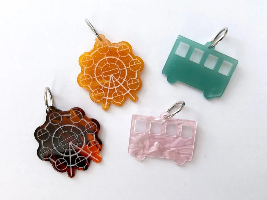白い背景に、観覧車とバスの形をしたカラフルなレジン製キーホルダーが4つ並べられています。オレンジのキラキラした観覧車、べっ甲柄の観覧車、ティールグリーンのバス、ピンクのマーブル模様のバスがあり、それぞれに銀色の金具が付いています。