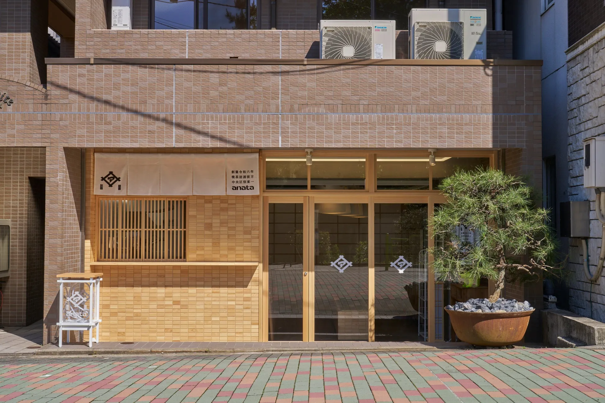 木材を基調とした洗練された外観の店舗。入り口にはガラス戸があり、店名が書かれた暖簾がかけられている。左側には格子状の窓、右側には盆栽が置かれている。