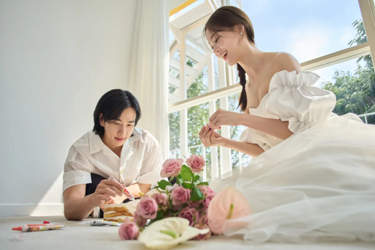 男性とウェディングドレスを着た女性が床に座ってケーキをデコレーションしている様子を描いた写真。ケーキの周りには花が飾られている。窓から明るい光が差し込んでいる。