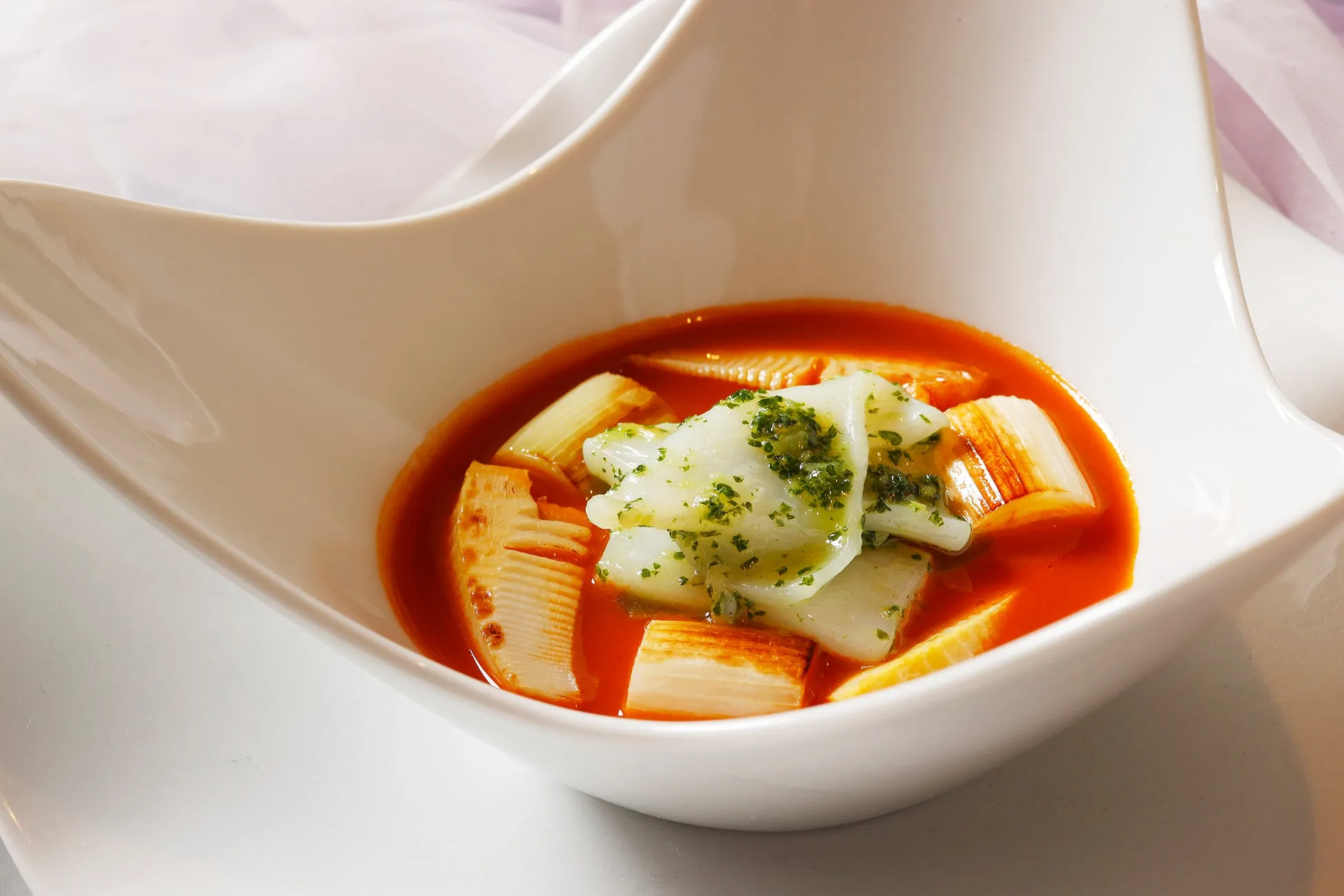 白い器に入った料理の写真。赤いスープの中に、白身魚のようなものと、ネギのような野菜、そしてハーブが乗っている。