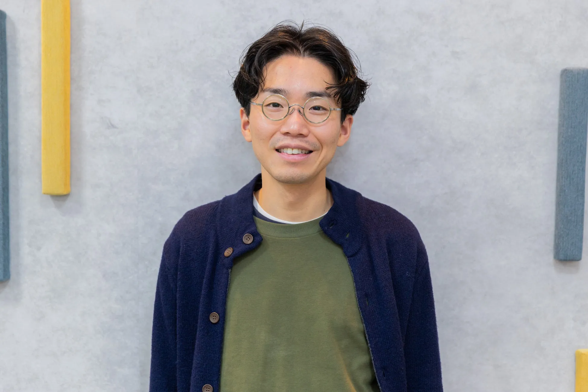 眼鏡をかけた若い男性が、カメラに向かって笑顔で立っています。彼は濃い青色のカーディガンと緑色のセーターを着ています。背景は灰色の壁で、黄色、青、緑の装飾的な要素がいくつかあります。