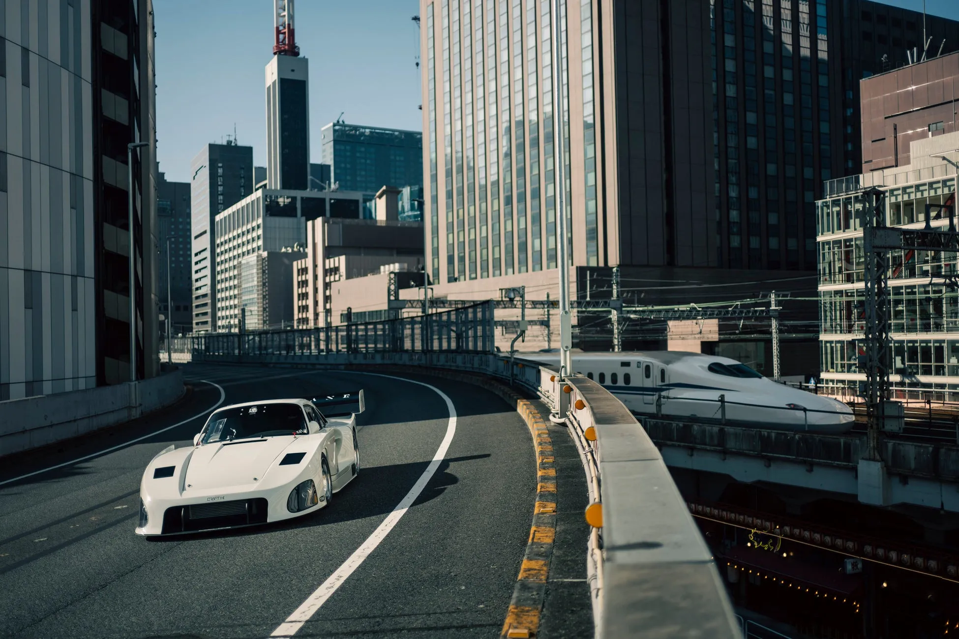 白いスポーツカーが街中を走っている様子を捉えた写真。背景には高層ビルや電車が映り、都会的な雰囲気を醸し出しています。