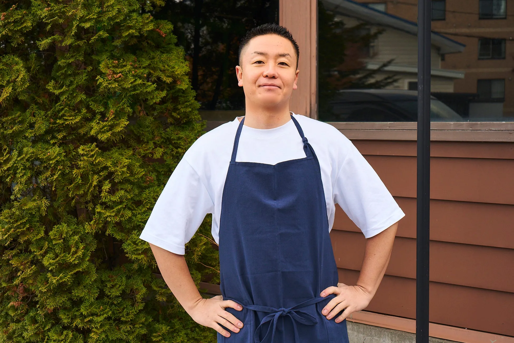 紺色のエプロンを着た男性が、屋外でポーズをとっています。彼は白いTシャツを着ており、彼の後ろには緑の植物と茶色の壁があります。