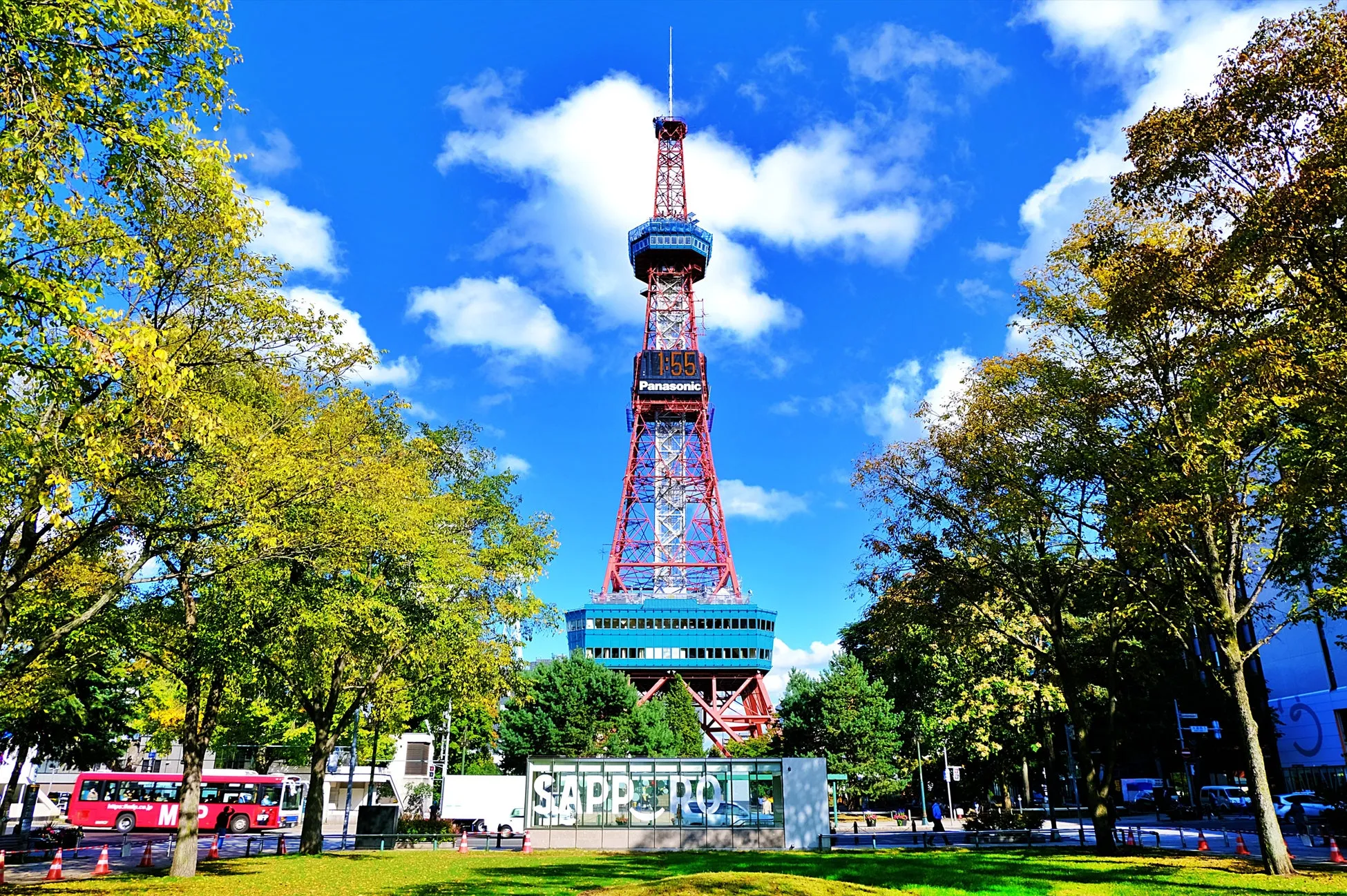 札幌テレビ塔の風景写真。青空の下、塔がそびえ立ち、周囲には緑豊かな木々が配置されています。手前には「Sapporo」の文字が見え、赤いバスも走っています。