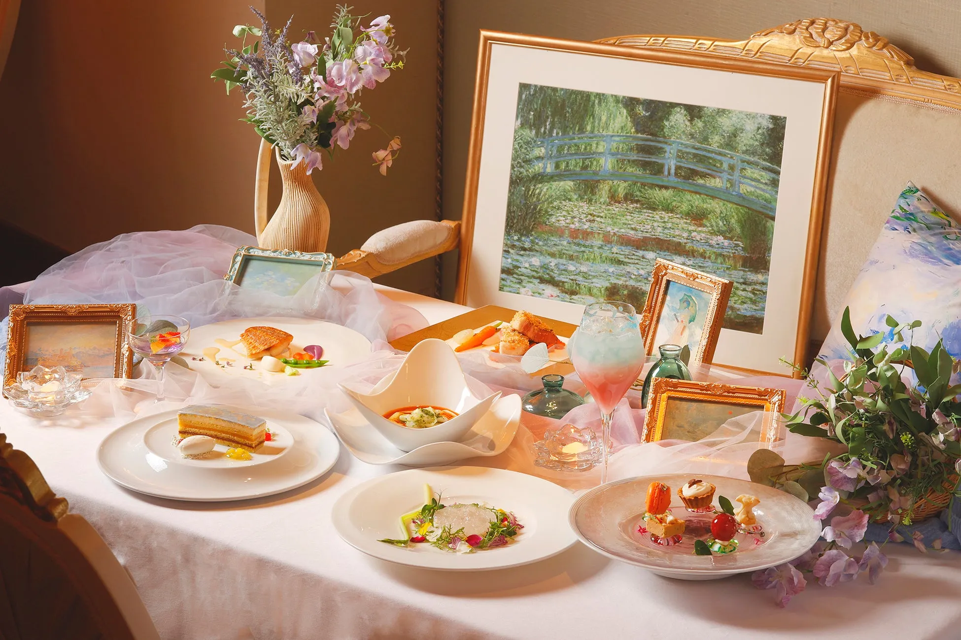 テーブルの上に、絵画、花、料理、飲み物などが美しく配置された、芸術的な雰囲気の画像。モネの絵画を思わせる水辺の風景が飾られ、それを取り囲むように、様々な料理が並べられている。料理は芸術作品のように盛り付けられ、テーブル全体が洗練された印象を与えている。