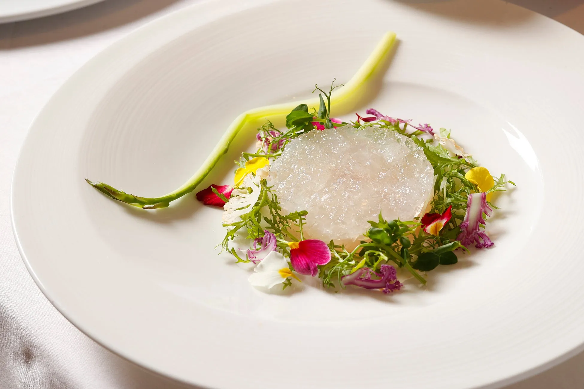 白い皿に盛り付けられた料理。中央に透明なゼリー状のものが置かれ、その周りを花やハーブで飾り付け、キュウリが添えられている。