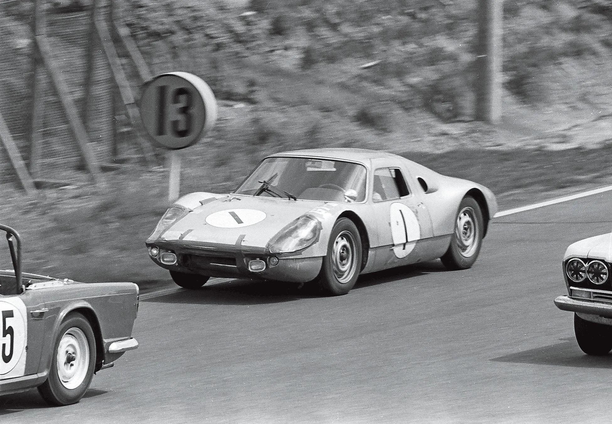 レース中のヴィンテージカーの白黒写真。ナンバー1の車がリードしており、ナンバー13の標識が背景に見える。