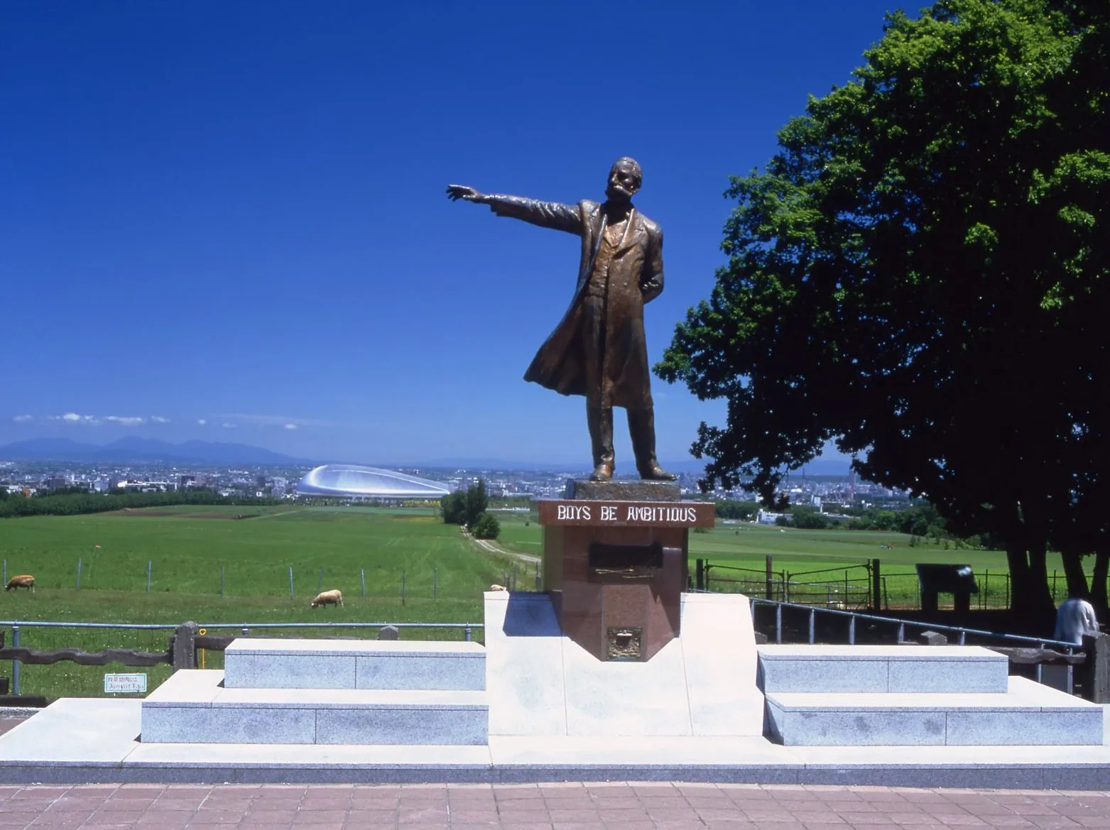 札幌の羊ヶ丘展望台にあるクラーク博士の像の写真。像は右手で遠方を指し、その下には「BOYS BE AMBITIOUS」という文字が刻まれている。背景には緑豊かな丘と札幌の街並み、青い空が広がっている。