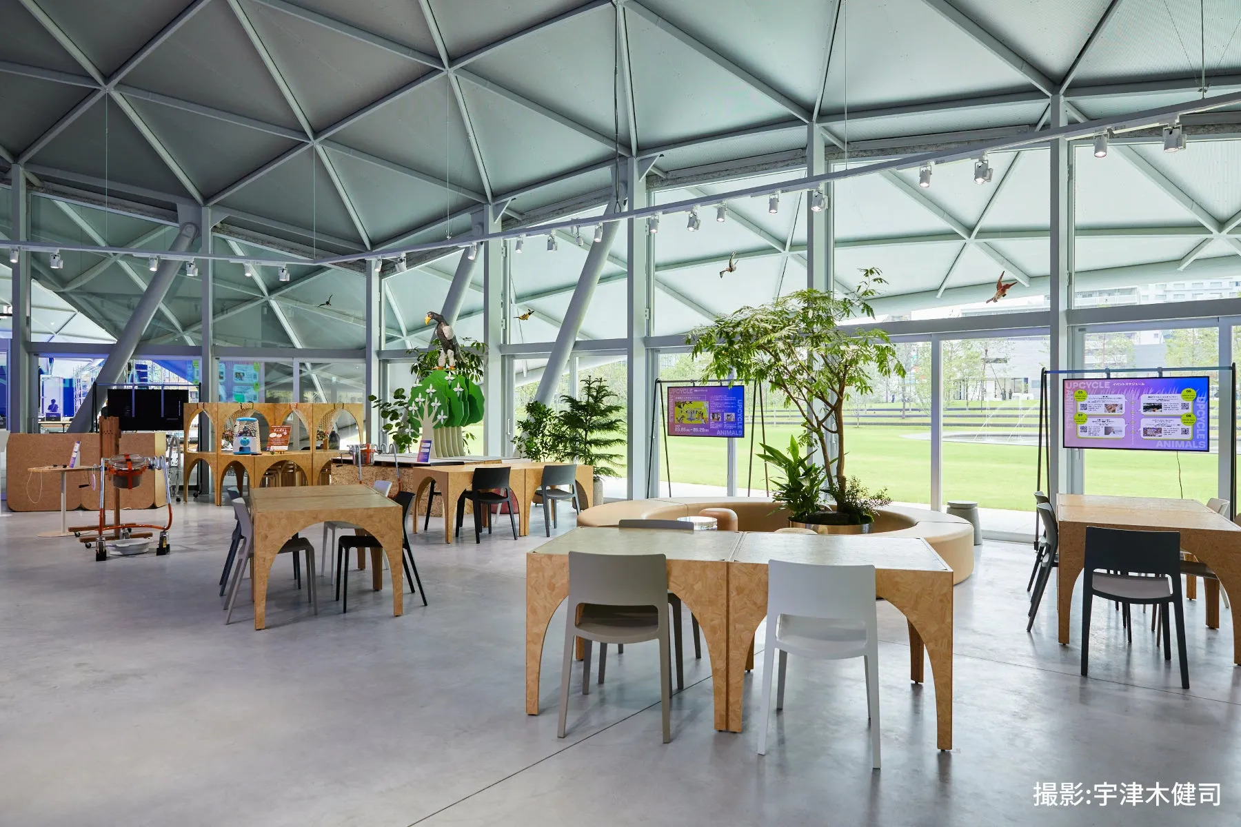 広々とした空間に、テーブルと椅子が配置され、窓からは緑豊かな景色が見える。天井は幾何学的な構造で、モダンな印象を与える。展示物やモニターが設置されており、イベントスペースや展示会場として利用されている様子。