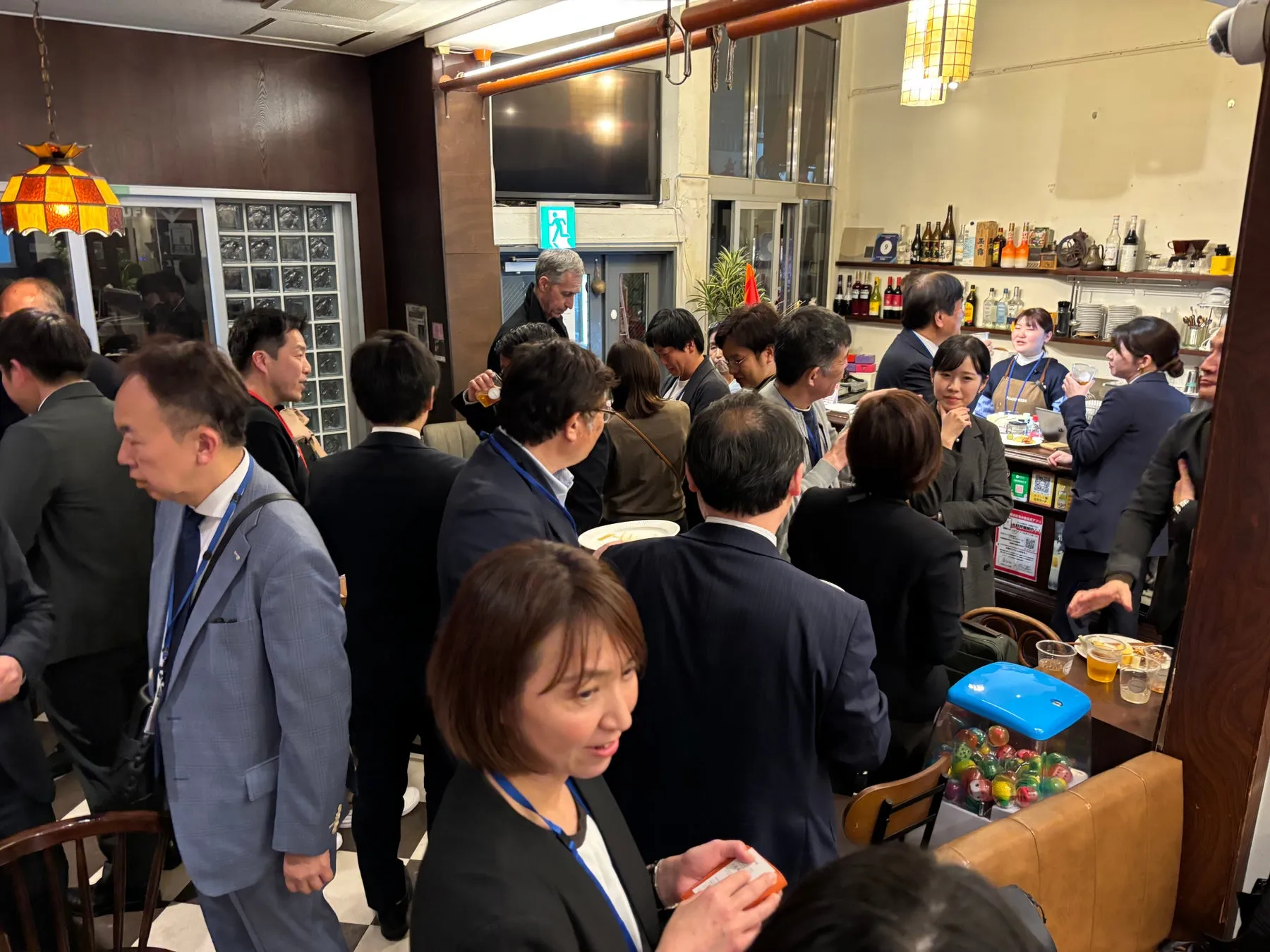 多くの人々が集まり、飲食をしている様子が写った写真です。バーのような雰囲気の場所で、人々は会話を楽しんだり、飲み物を手にしています。