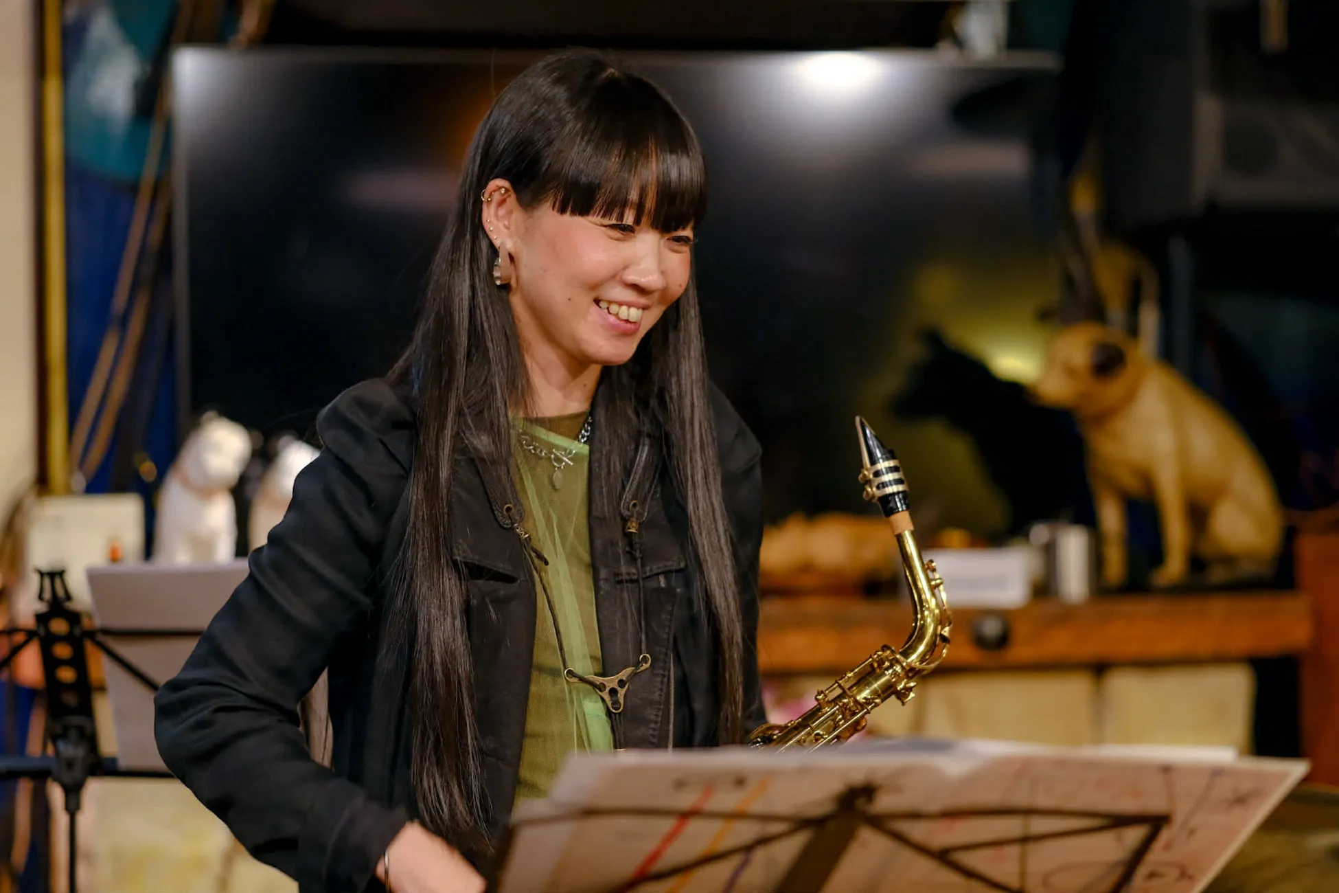 サックスを演奏する女性の笑顔のポートレート。彼女は黒いジャケットと緑色のインナーを着ており、長い黒髪をしている。背景には、犬の置物、楽譜、音楽スタンドがある。