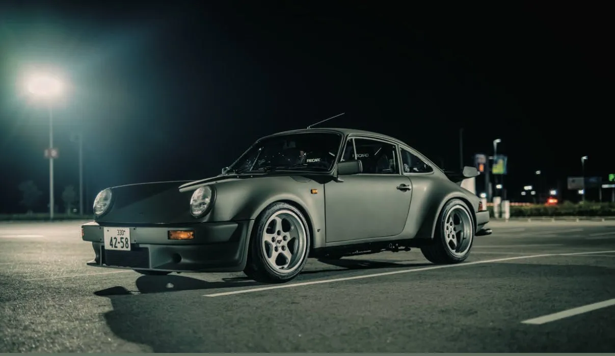 夜の駐車場に駐車された、グレーのポルシェ911の画像です。車は左側から撮影されており、ヘッドライトが点灯しています。車のナンバープレートには数字と文字が記載されており、座席には「RECARO」のロゴが見えます。