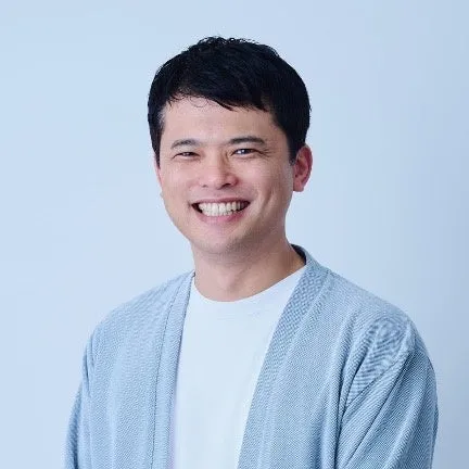 笑顔の男性のポートレートです。彼は白いTシャツと水色のカーディガンを着ています。背景は白です。