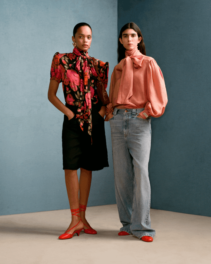 2人の女性が立っており、それぞれ異なるファッションを着ている。左の女性は赤い花のプリントのブラウスと黒いスカートを着ており、赤い靴を履いている。右の女性はピンクのブラウスとジーンズを着ており、赤い靴を履いている。