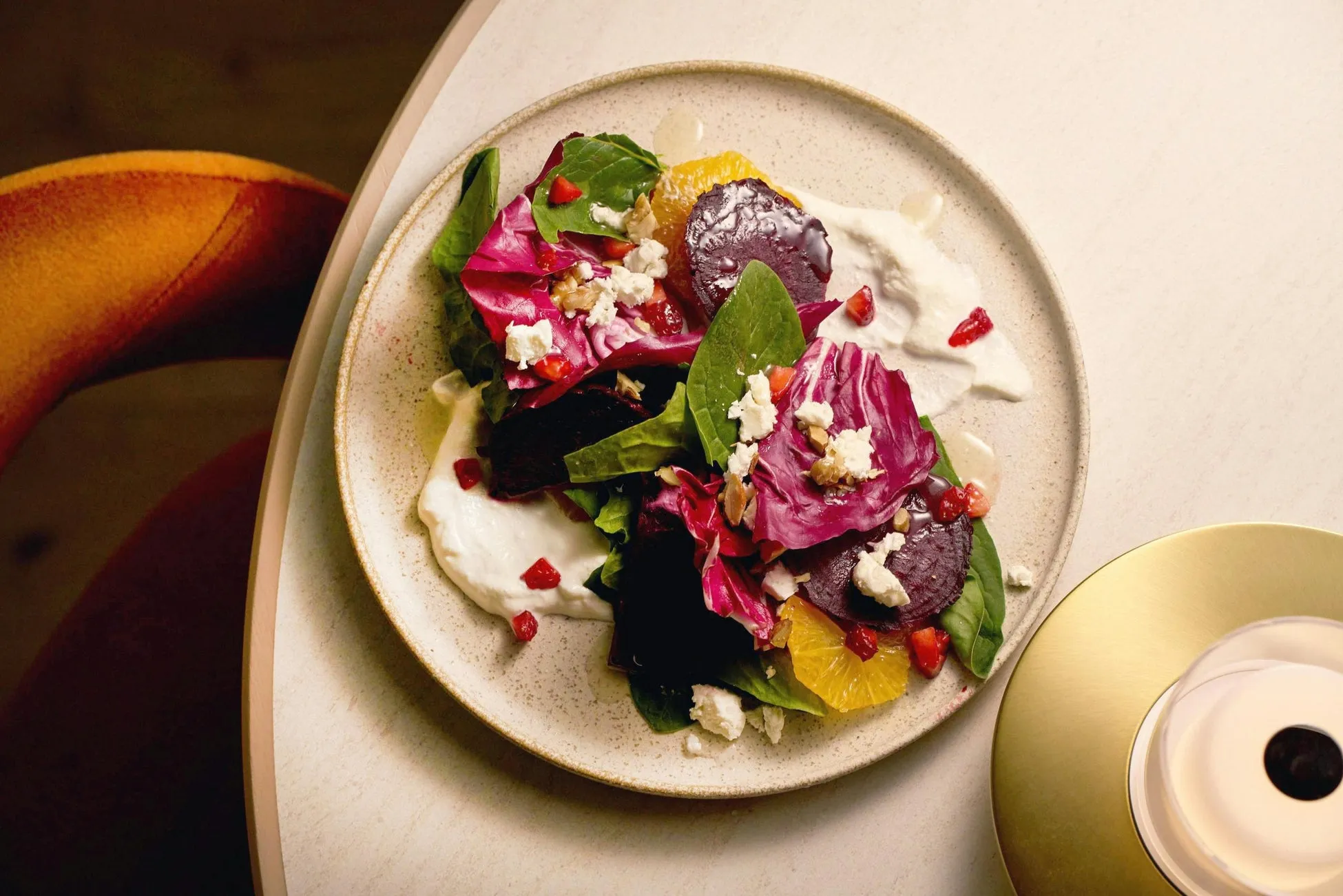 お皿に盛り付けられたサラダのクローズアップ写真。サラダは、ビーツ、オレンジ、葉野菜、チーズ、ザクロで構成されている。サラダは白いソースの上に配置され、テーブルの上に置かれている。