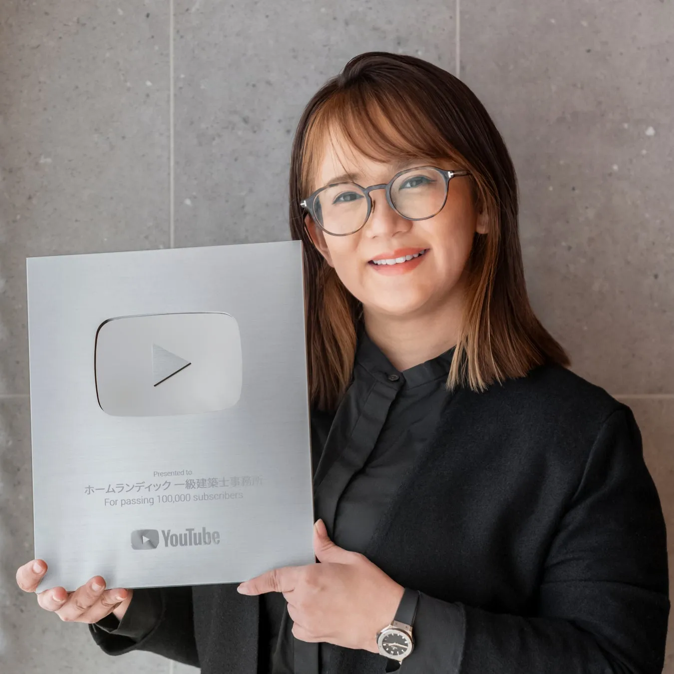 女性がYouTubeの10万人の登録者達成を記念した銀色のプレートを持っています。プレートには「ホームランディック一級建築士事務所」と書かれています。