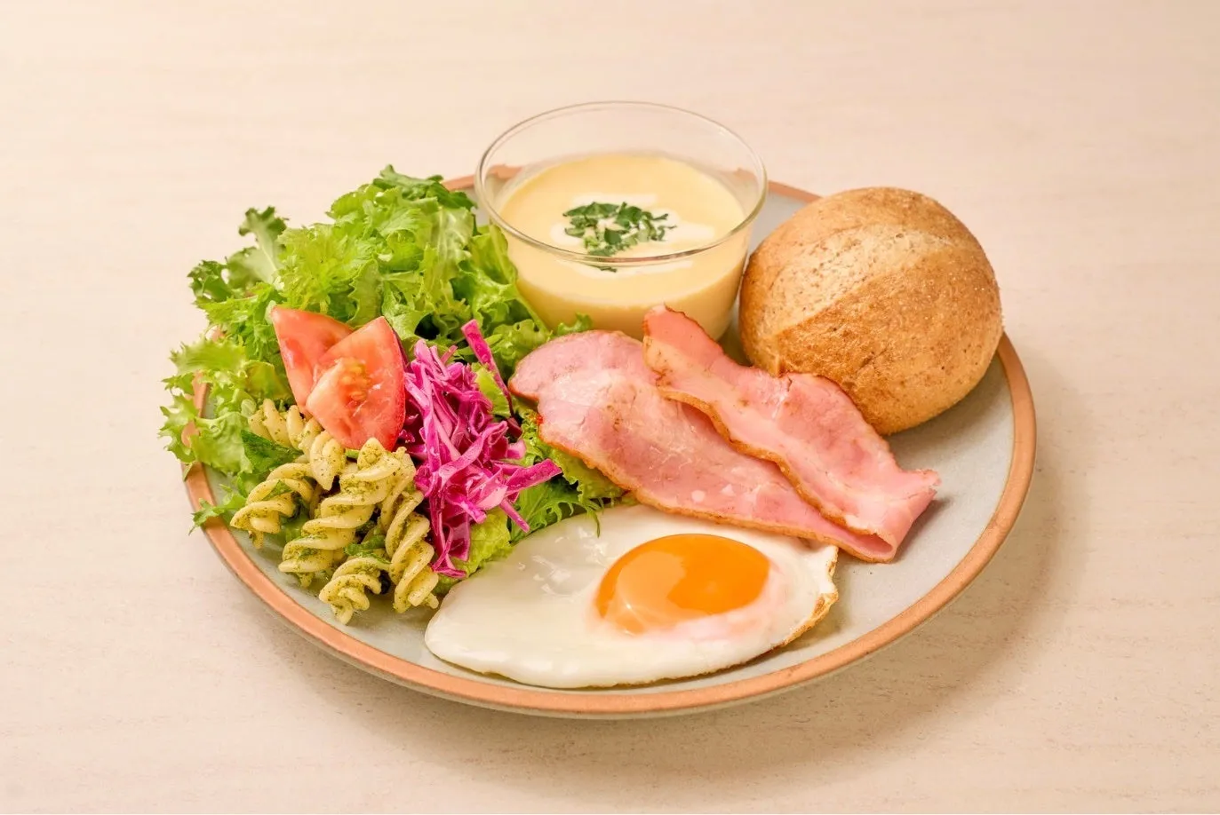 朝食のプレート。サラダ、パスタ、トマト、紫キャベツ、ベーコン、目玉焼き、パン、スープが乗っています。