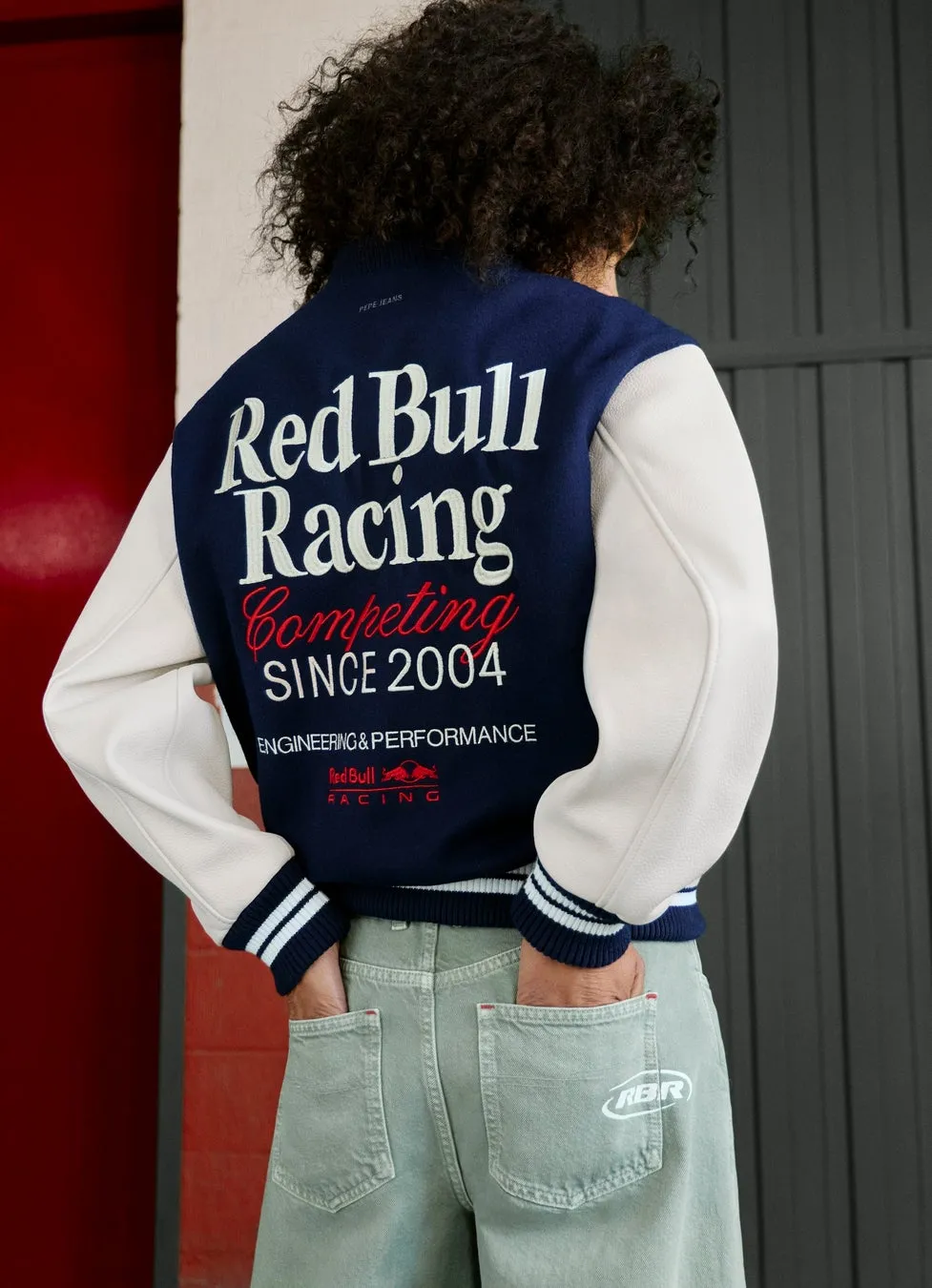 後ろ姿の男性が、背中に「Red Bull Racing」と書かれたスタジアムジャンパーを着ています。彼は、緑色のジーンズを履いており、ポケットに手を入れています。