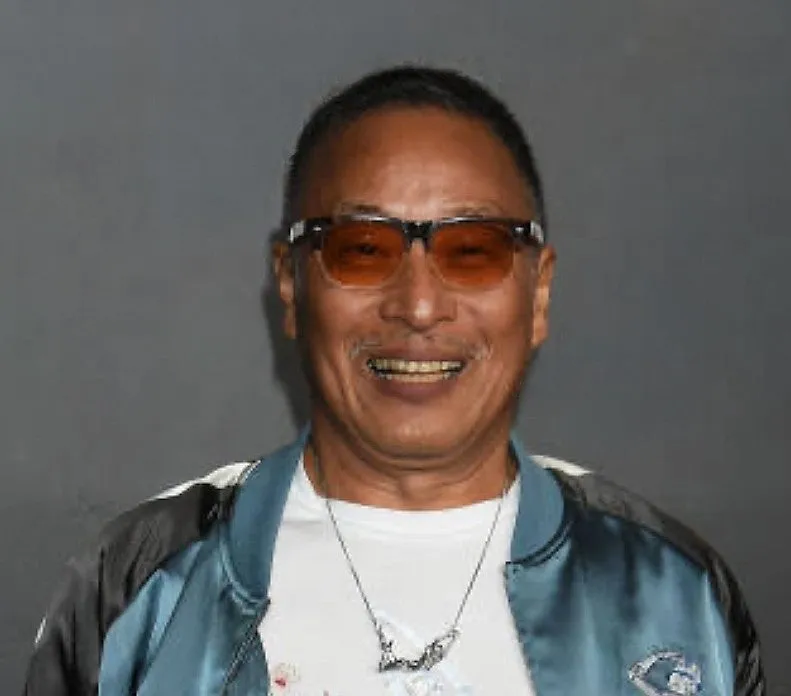 サングラスをかけた男性のポートレート。彼は白いTシャツを着ており、青いジャケットとネックレスを身につけています。背景は単色です。