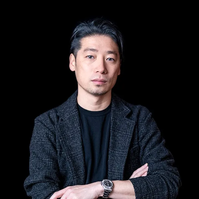 黒い背景に、腕を組んでこちらを見つめる男性のポートレート。