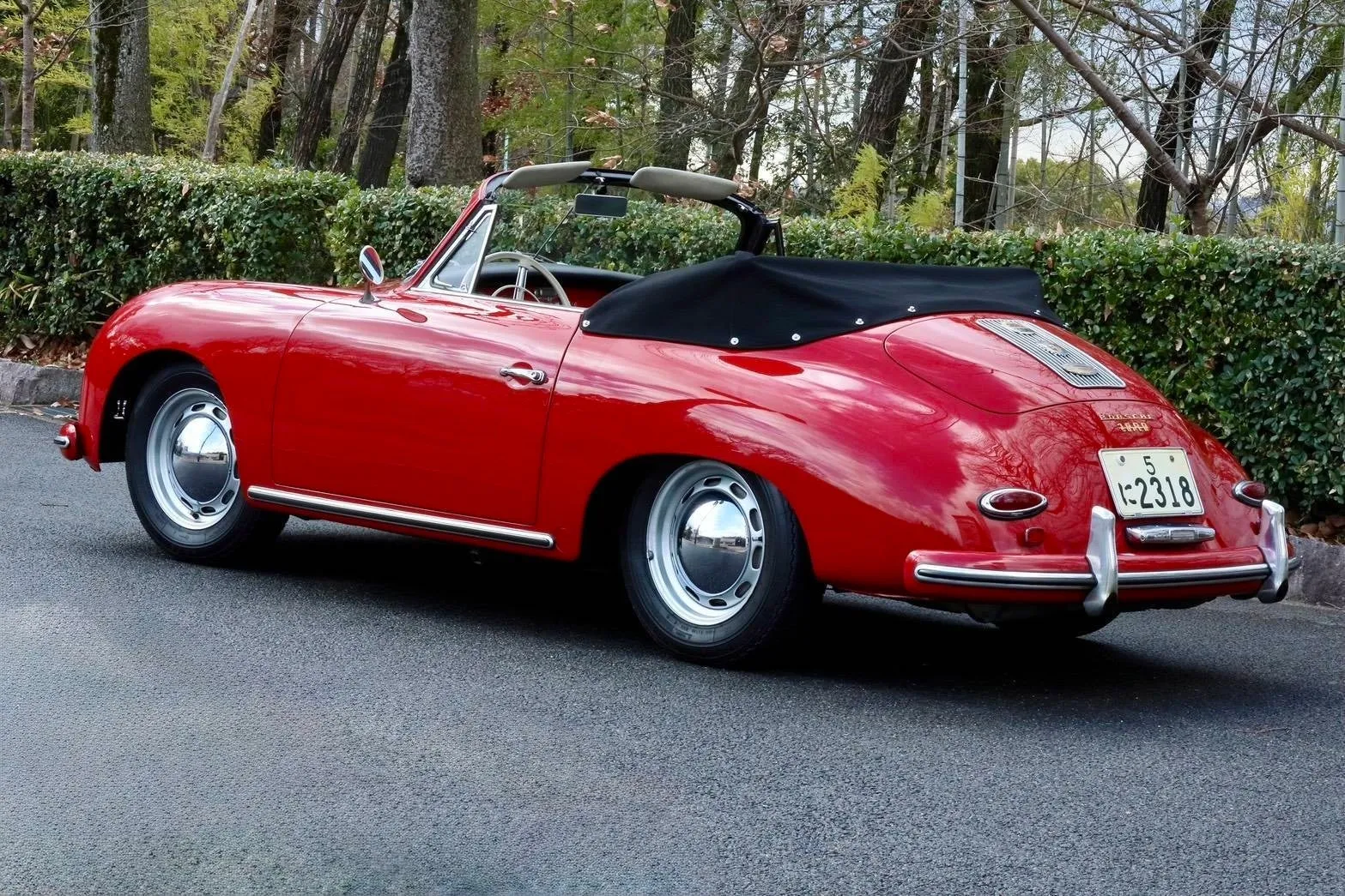 赤いポルシェ356のコンバーチブルが、アスファルトの道に駐車されています。車の背面にはナンバープレートが付いており、背景には緑の生け垣と木々があります。