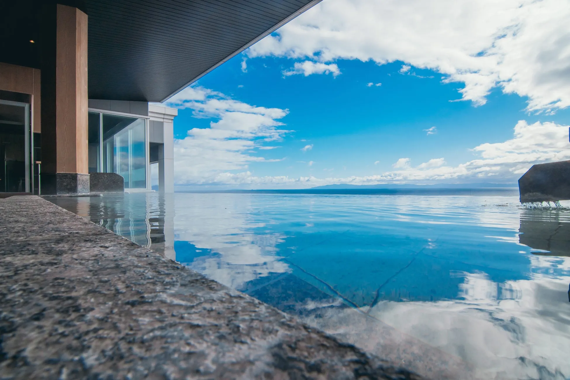 青い空と雲が水面に反射する屋外プール。建物の一部が写り込んでおり、開放感のある空間が広がる。