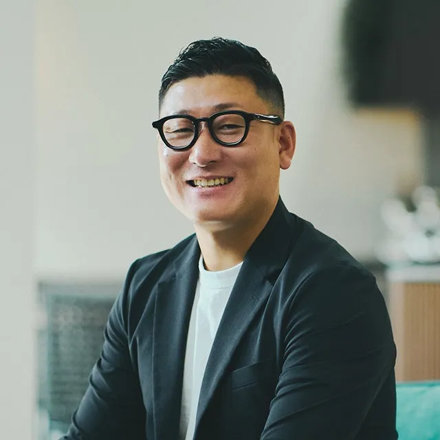 眼鏡をかけた男性が笑顔でカメラを見ているポートレート写真です。男性は黒いジャケットと白いTシャツを着ています。