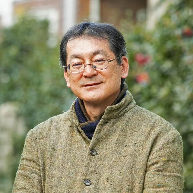 眼鏡をかけた、笑顔の日本人男性のポートレートです。彼はカメラのほうを見ています。背景にはぼやけた緑色の植物が見えます。