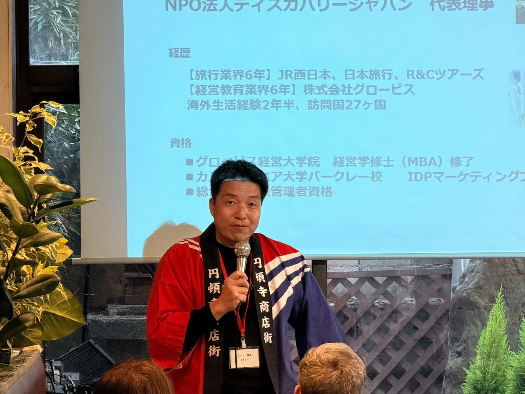 プレゼンテーションをしている男性の画像。背景には、彼の経歴や資格が書かれたスライドがある。