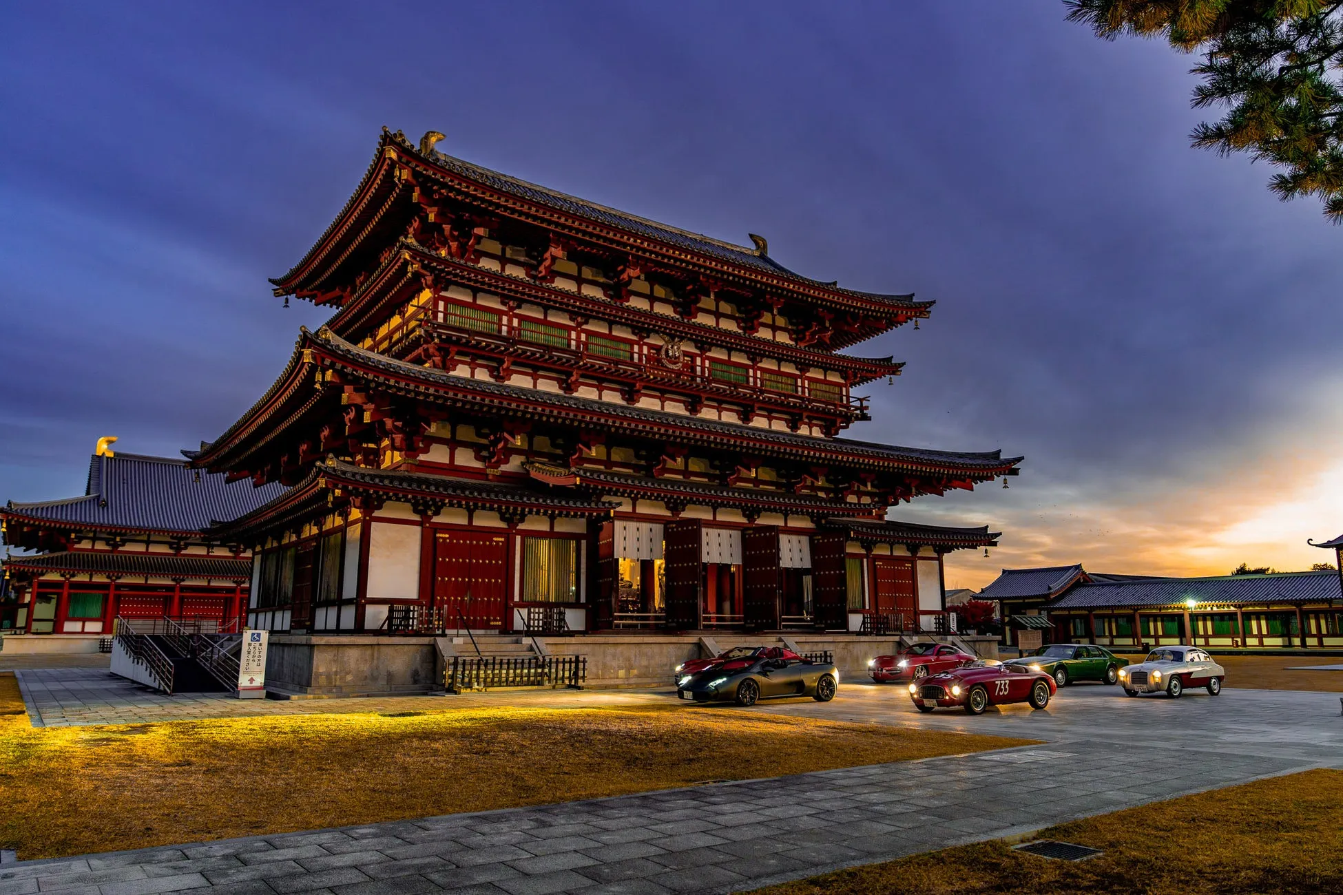日本の寺院の前にクラシックカーが並んでいる様子を描いた写真。空は夕焼けで、建物のライトアップが美しい。